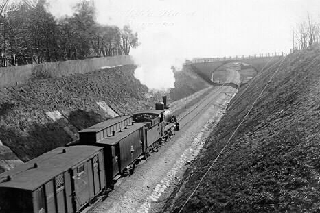 Gammalt svartvitt foto på godståg med ånglok som kör mot en välvd järnvägsbro.