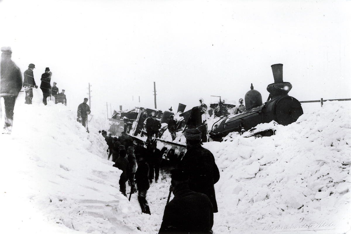 Ett gammalt ånglok har spårat ur och sitter fast i en snödriva. Människor står och tittar på, några gräver i snön med spadar. Svartvit bild från början av 1900-talet.