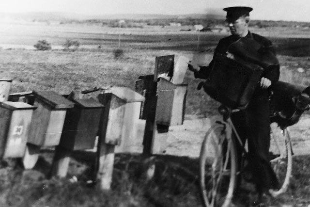 Cyklande man i uniform lägger ett brev i en brevlåda. Svartvit bild från 30-talet.