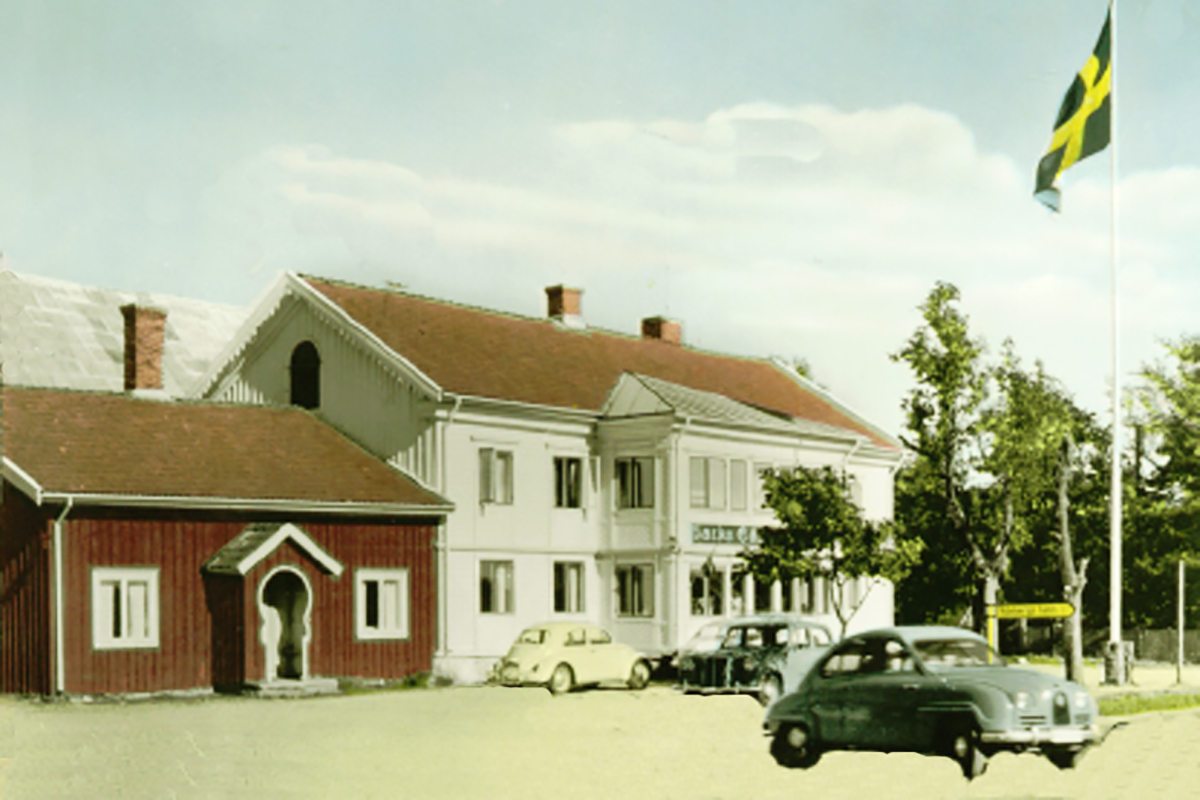 Gästgiveriet i Väröbacka. En gammal röd träbyggnad från 1600-talet sitter ihop med en vit träbyggnad från 1700-talet. Framför står tre bulliga bilar från 60-talet. Flaggan vajar i topp på en flaggstång.