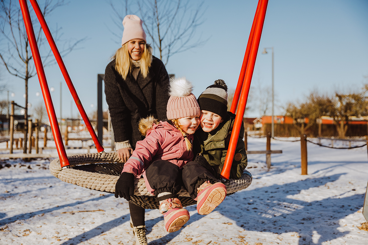 Kvinna gungar två barn på en vintrig lekplats.